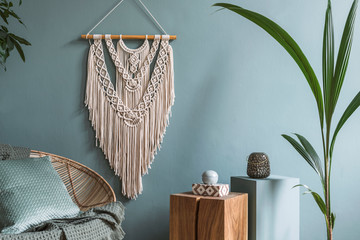 Interior design of living room at modern home with stylish rattan armchair, pillows, plaid, beige macrame, wooden cubes, tropical plants and elegant accessories. Design home decor. Eucalyptus walls. © FollowTheFlow