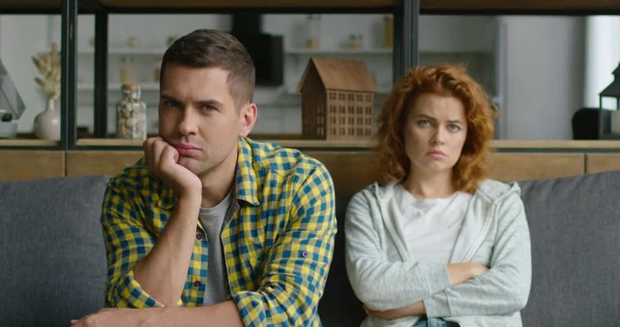 Portrait of young man and woman, sad and unhappy, looking at camera, woman is blurred, feeling guilty for past, indoor, struggle in relationships, sitting on couch at home. 4K, shot on RED camera.