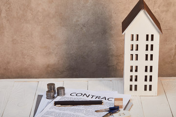 house model on white wooden table with contract, coins and keys near brown wall, real estate concept