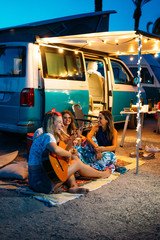 Group of happy people playing the guitar together and chatting near camper van, after dinner. Holiday and friendship concept. - obrazy, fototapety, plakaty