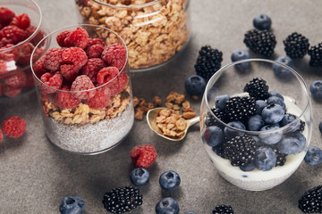 selective focus of tasty yogurt with chia seeds, berries and oat flakes in glasses near teaspoon on marble surface