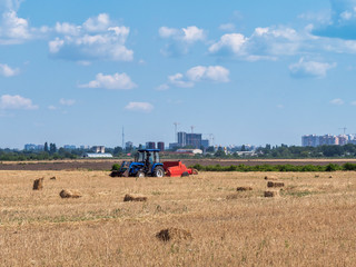 Fototapeta premium Hay is packaged in briquettes on a field under a blue sky with clouds. Works special agricultural equipment. The village and the city . In the background is the construction of the city. Urbanization.