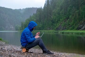 The guy in the blue jacket with a hood with a laptop in his lap sits on the bank of the river in...