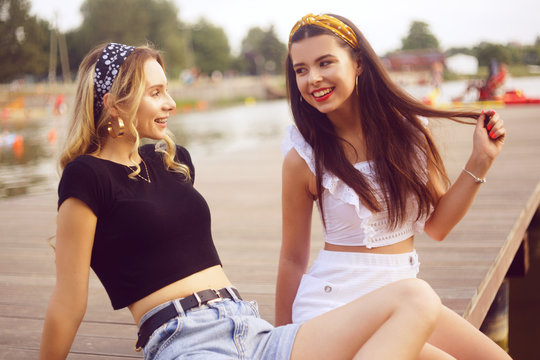 Two fashionable girls, girlfriends, are sitting on the pier near the river, the beach. Girls talk on the beach, laugh, smile, share secrets, discuss guys. Rest in the summer, a trip with a friend.