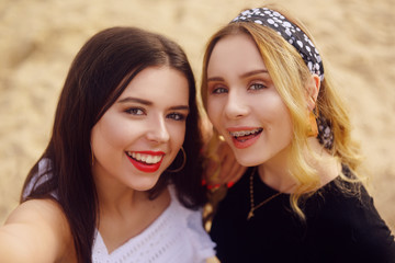 Fototapeta premium Girls on the beach stand on the background of sand. The brunette in white and the blonde with a bandana on her head. Beautiful girls look at the camera and take a selfie. Girls make photos on camera.