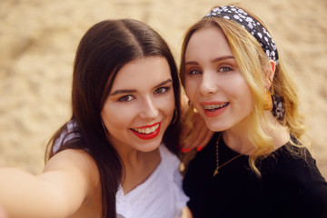 Girls on the beach stand on the background of sand. The brunette in white and the blonde with a bandana on her head. Beautiful girls look at the camera and take a selfie. Girls make photos on camera.