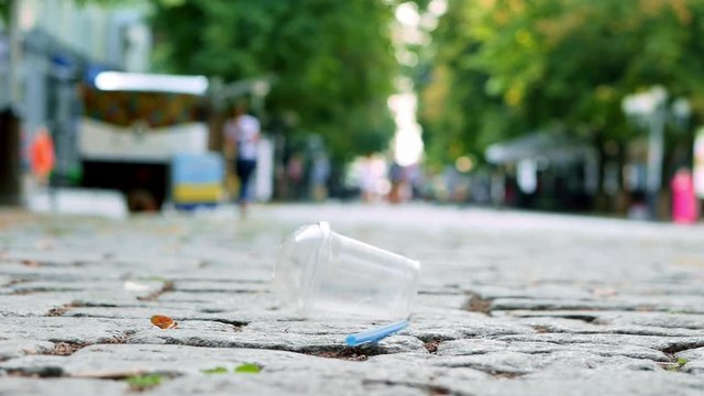 Plastic pollution, plastic cup falls down on the city street in slow motion, coffee shop and people in the background