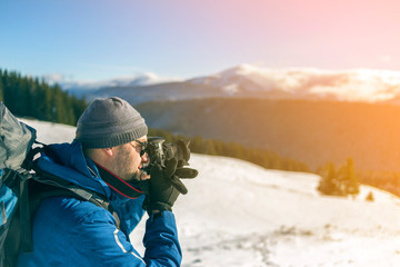 Hiker man tourist photographer in warm clothing with backpack and camera taking picture of snowy valley and woody mountain peaks landscape under blue sky on sunny winter cold day.