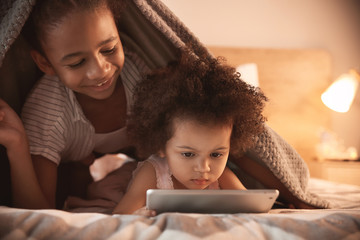 Cute African-American sisters with tablet computer watching cartoons in bedroom