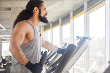 Closeup side view portrait of young adult sport man with long curly hair training alone. Handsome male doing exersices in a gym on elliptical machine and looking at window. Indoor, fitness concept