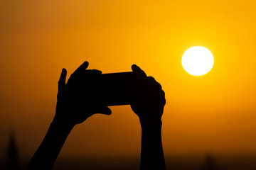 Silhouette of hands holding a mobile phone and making photography of the huge sun during sunset or sunrise. Male makes a photo on his phone.