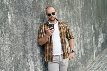 waist up portrait of young good looking bold bearded guy standing outdoors against grey modern loft wall with call phone in his hand chatting. wearing yellow shirt, jeans and sunglasses.