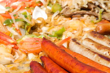 Hot dogs and sausages with onions, jalapenos, and several varieties of bell peppers and tomatoes grilling on an outdoor vendors grill. Close up.