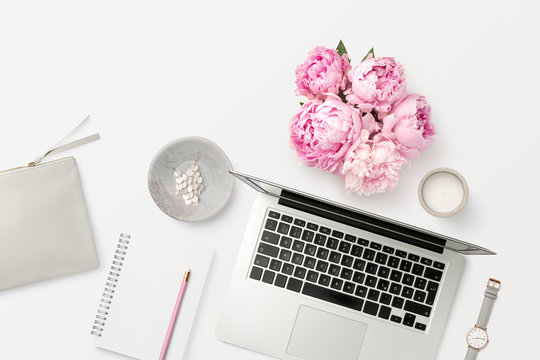 bright feminine workspace with open laptop computer, office supplies, fresh flowers, stylish clutch bag and other accessories on a white desk, top view / flat lay, copyspace for your text