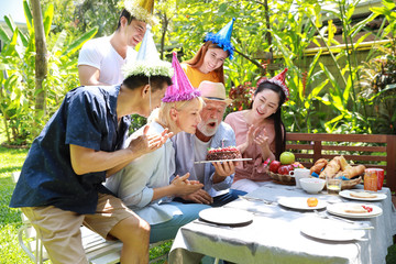 Happy multiethnic family giving surprise gift to caucasian grandfather on his happy birthday and he blowing out candles on homemade baked cake with happy face in backyard outdoor on sunny day