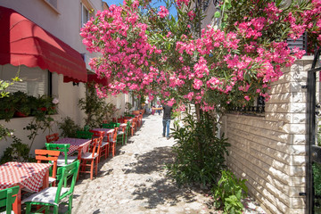 Cafe tables and chairs and pink flowers in the street. People walking on the street.