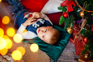 Fototapeta na wymiar a little boy is lying on the floor on a pillow near a Christmas tree with bokeh lights garlands
