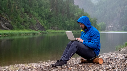 A man in a blue jacket with a hood is working on a white laptop while sitting on a log in the rain by the river. Programmer or Hacker in the mountains. Concept waterproof computer equipment.