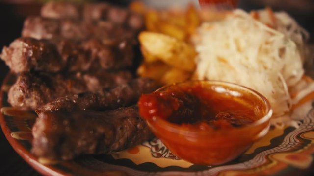 A fork picks up a grilled Balkan cevapi sausage, dips it into a traditional sauce, and a person takes a bite off-camera in a rustic restaurant.