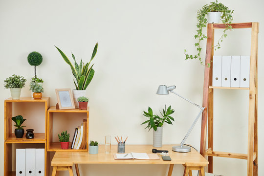 Image of modern workplace with wooden table and notepad on it and different kinds of green plants on the shelves