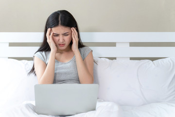 Young beautiful Asian woman sitting on bed, She use laptop  feel serious and headache