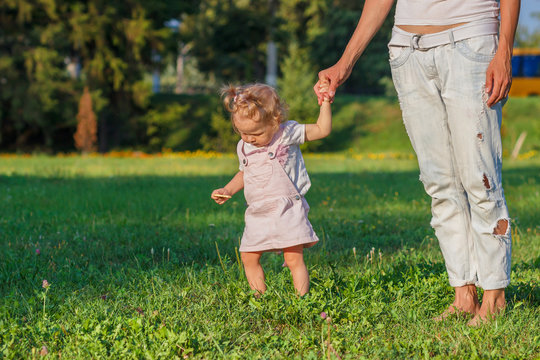 Little cute girl does her first steps with mother support, green grass, summer time, blurred background