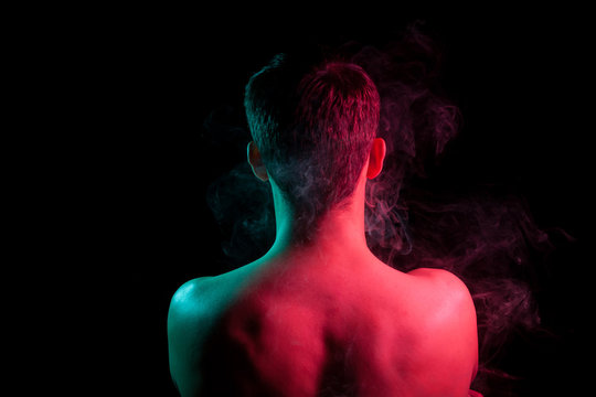 Close-up of the nape, back and shoulders of an short hair head of a man with stubble, naked without clothes, illuminated on one side in red and on other green on a black isolated background with smoke