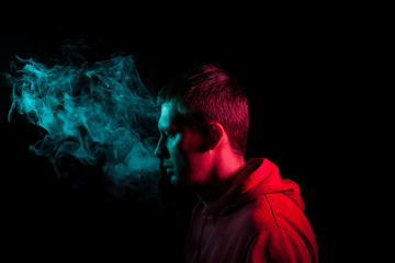 Close up portrait of the face of an adult serious man exhales green toxic smoke while smoking e-cigarette and vape illuminated with blue and red colored light on a black background. Harm to health.