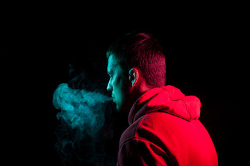 Close up portrait of the face of an adult serious man exhales green toxic smoke while smoking e-cigarette and vape illuminated with blue and red colored light on a black background. Harm to health.