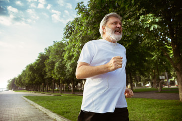 Senior man as runner with armband or fitness tracker at the city's street. Caucasian male model practicing jogging and cardio trainings in summer's morning. Healthy lifestyle, sport, activity concept.