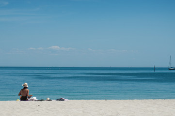 Portrait of woman in swimsuit and hat sitting on the beach on back view with sailboat on background