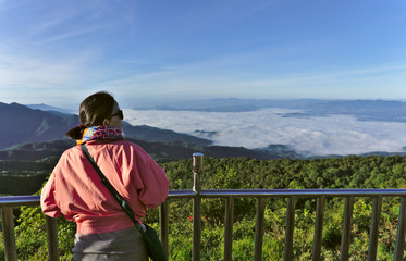 Fototapeta na wymiar Female asian traveller with black hair wearing pink jacket and glasses holding stainless steel fence looking to the scenic view of fog, mist, forest and mountain at Doi inthanon Chiangmai Thailand