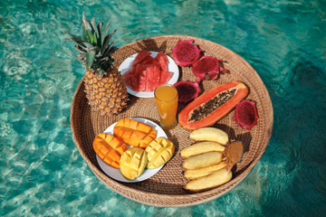 Basket with tropical fruits ( pineapple, banana, papaya, pitahaya, manho juice) floating in the pool. Cocept healthy food, floating breakfast tray in the swimming pool. Summer vacation on Bali