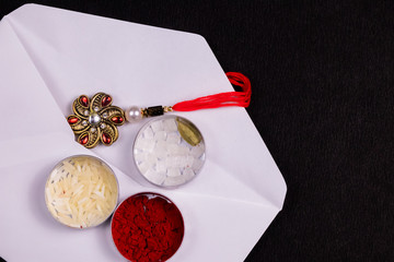 Indian Festival concept - Elegant Rakhi in a white envelope with rice grains, kumkum, sugar rock and cardamom a flat tin box on black background