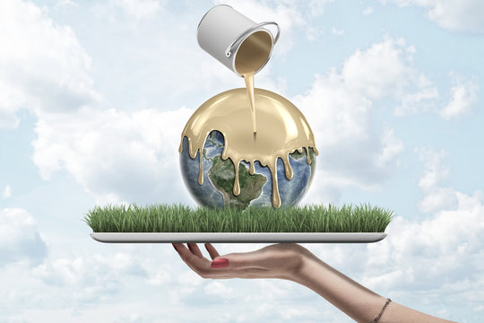 Closeup of woman's hand facing up holding digital tablet with Earth lying on screen and paint can in air pouring gold paint onto Earth.
