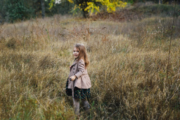 fair-haired girl in a beige coat on the dry grass and autumn trees background.