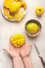 Mango preparation step by step. Female hands holding half diced mango on white background with coconut bowl and fruits plate, top view.
