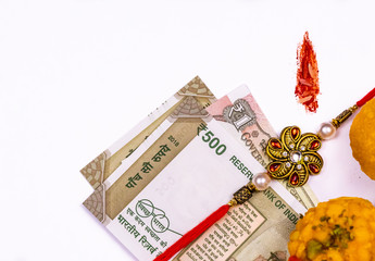 Raksha Bandhan concept - Elegant Rakhi with Indian currency, sweets and kumkum rice grain mark on white surface. Rakhi, Indian brother and sister festival