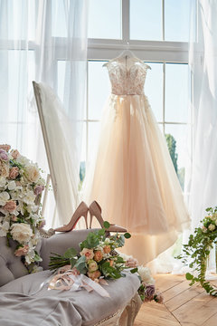Beautiful beige women's shoes and bridal bouquet on chesterfield sofa with wedding dress in blur hanging on hanger against window in hotel room, copy space