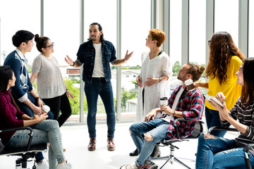 Attractive young multiethnic group talking at relax zone in coffee break time at office. Young asian hipster man sharing lifestyle idea to staff friends with fun and happy in happy workplace concept. - obrazy, fototapety, plakaty