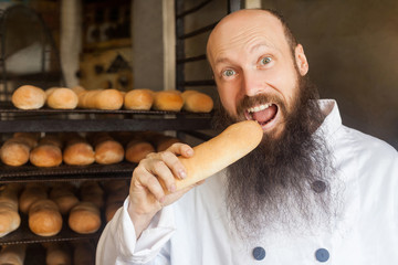 Portrait of happy young adult hungry baker with long beard in white uniform standing in his workplace and tasty fresh baked long loaf, Indoor, looking at camera, profession concept