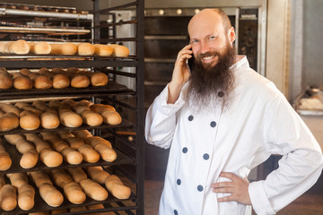 Portrait of happy young adult businessman baker with long beard in white uniform standing in bakery and have online order by phone, talking on cellphone with toothy smile Indoor, looking at camera