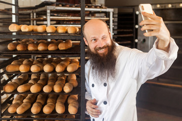 Portrait of cheerful young adult blogger baker with long beard in white uniform standing in factory and making selfie on shelves with fresh bread background, winking, Indoor, profession concept