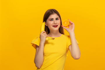 Young model in a yellow tee holds banana and orange standing over yellow background.