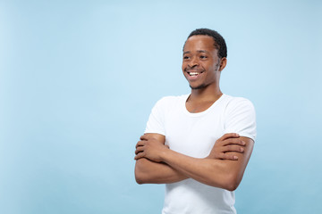 Half-length close up portrait of young african-american man in white shirt on blue background. Human emotions, facial expression, ad, sales, concept. Standing hands crossed and smiling.