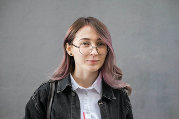 A young woman in a jeans jacket, eyeglasses. Hipster with purple colored head, in round glasses on a gray wall background.