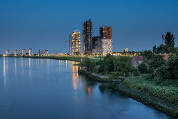 Fototapeta na wymiar luminated tall apartment buildings at the riverside after sunset. Spijkenisse skyline at the Oude Maas river. With small house surrounded by green and wave breakers rocks at the shoreline