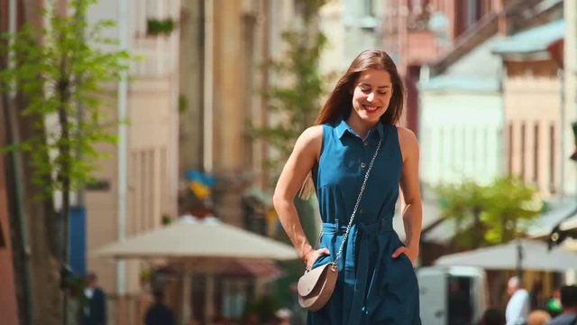 Charming young woman walking like model through city center on sunny day. Portrait of excited cheerful girl in blue dress hands in pockets enjoying sunlight and life. Beautiful cityscape.