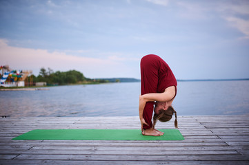 Beautiful young woman practices yoga asana Uttanasana - Standing Forward Fold on the wooden deck near the lake