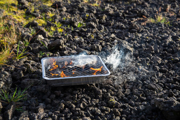 Obraz premium Small disposable barbecue stands on the stones. Flames come from charcoals of grill. Preparing BBQ, Iceland.